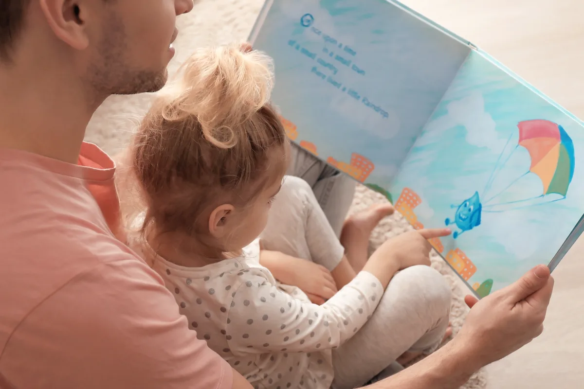 A parent reading a picture book aloud with a small child on their lap, the child's finger on the page