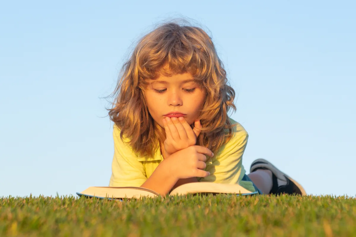 A child lying in the grass in the late sun, chin resting on their hands, lost in an open book